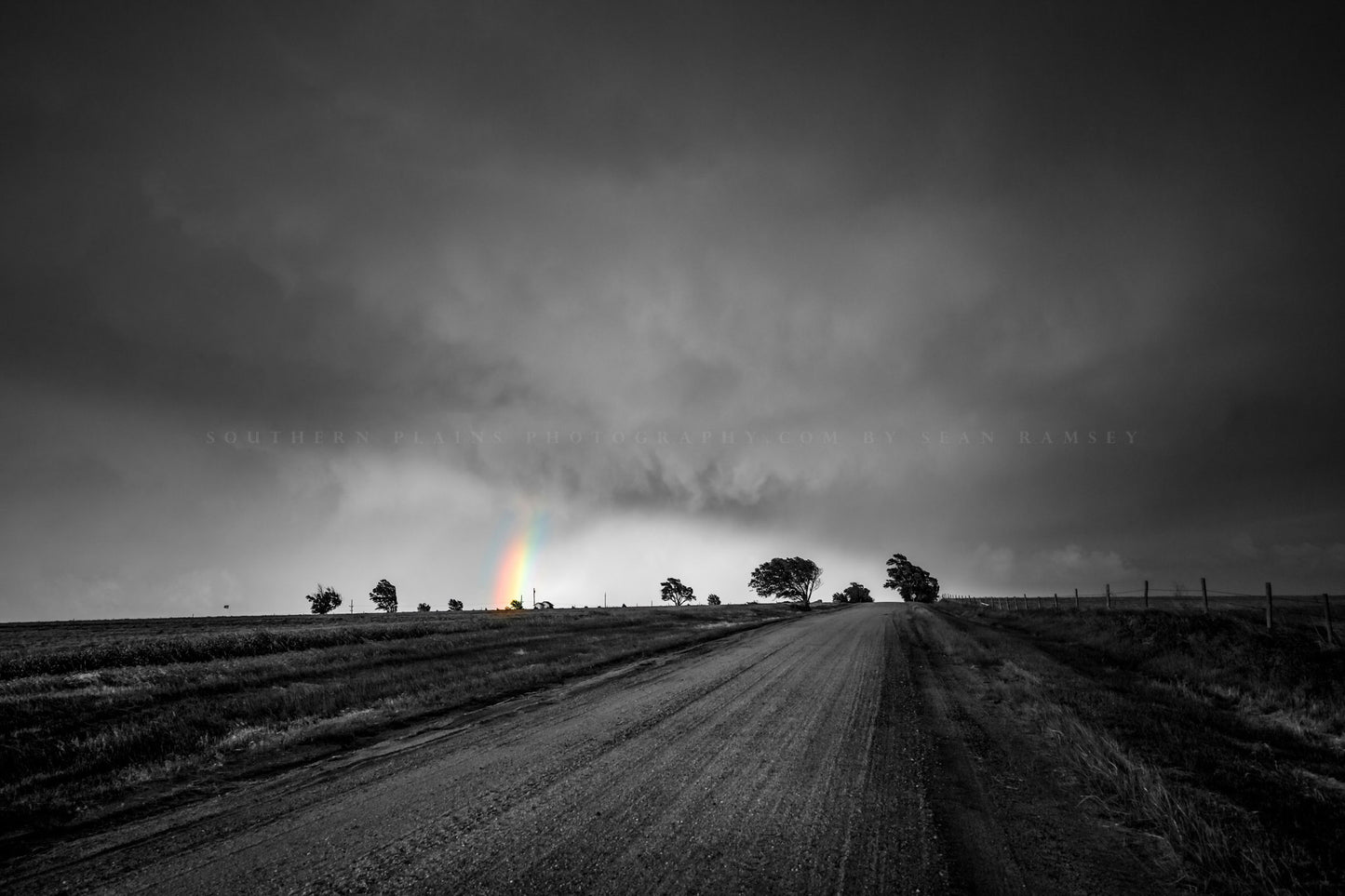 Moody Great Plains photography print of a colorful rainbow appearing under storm clouds down a dirt road in Kansas in black and white by Sean Ramsey of Southern Plains Photography. 