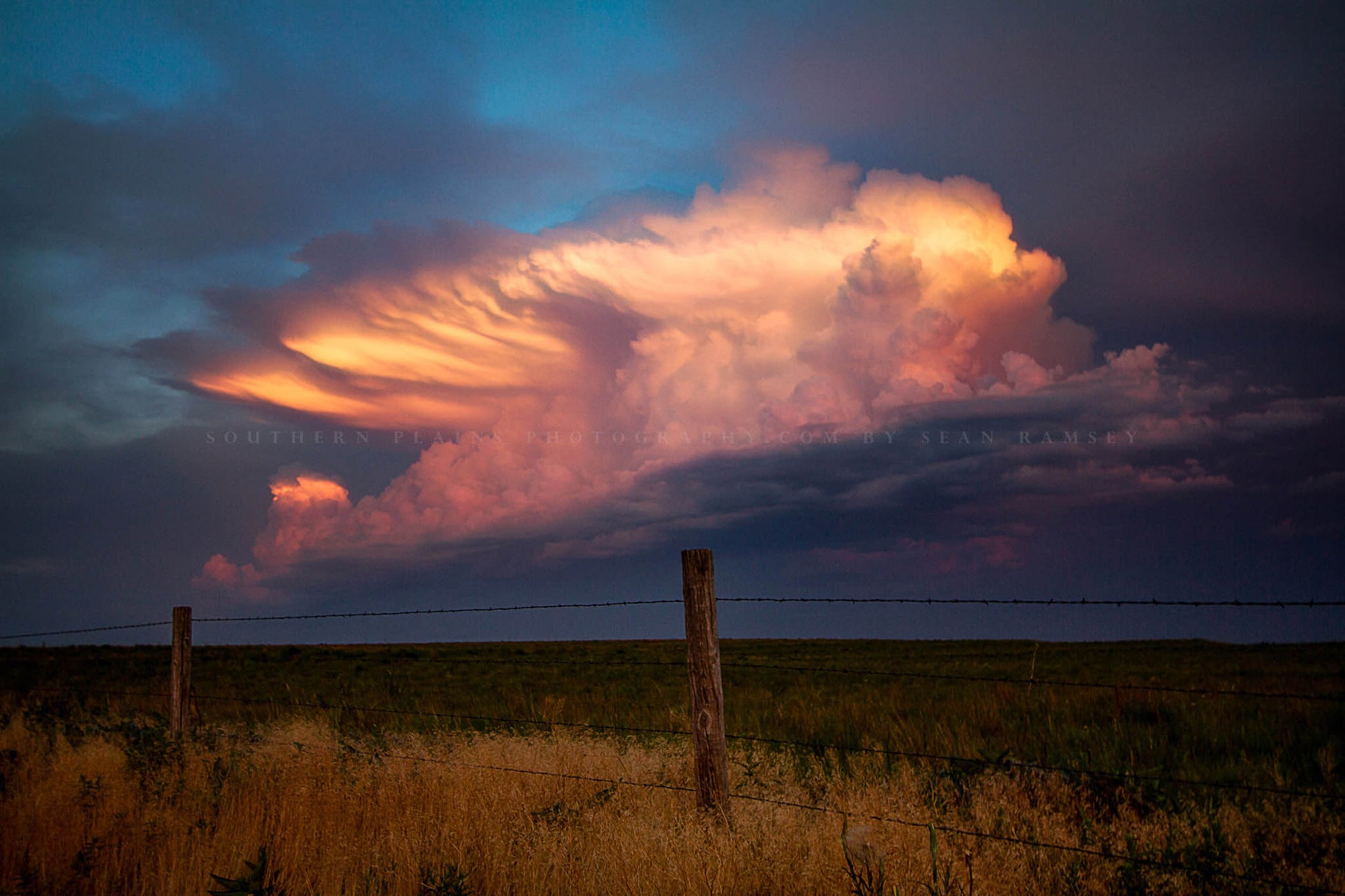 Storm photography print of a distant supercell thunderstorm drenched in sunlight over a barbed wire fence at sunset on a spring evening in Oklahoma by Sean Ramsey of Southern Plains Photography.