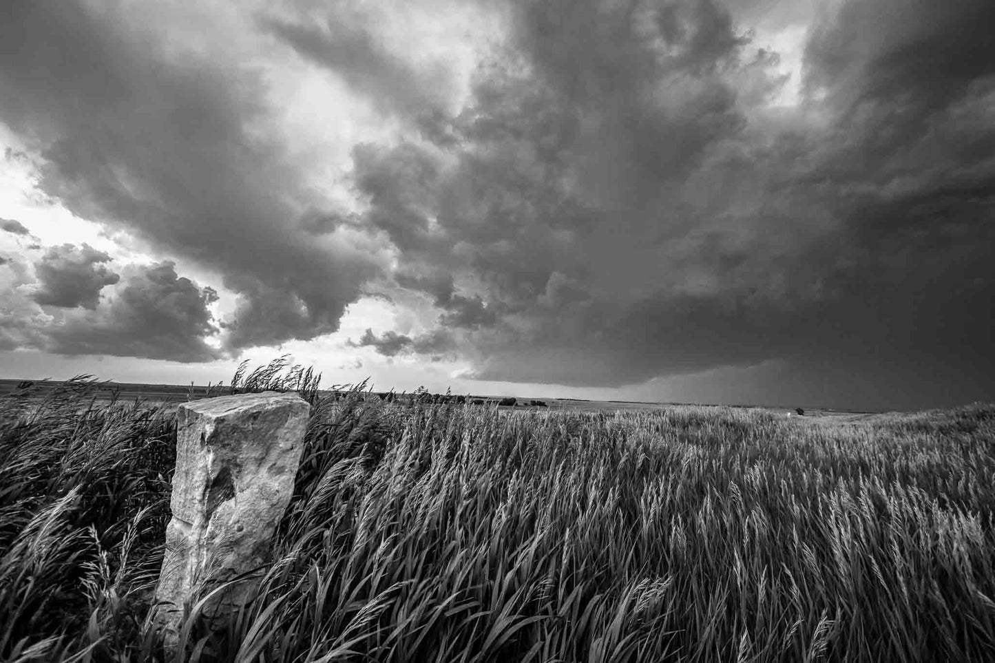 Black and white Great Plains photography print of an old limestone marker in waving prairie grass as a storm brews on the horizon on a stormy spring day in Kansas by Sean Ramsey of Southern Plains Photography.