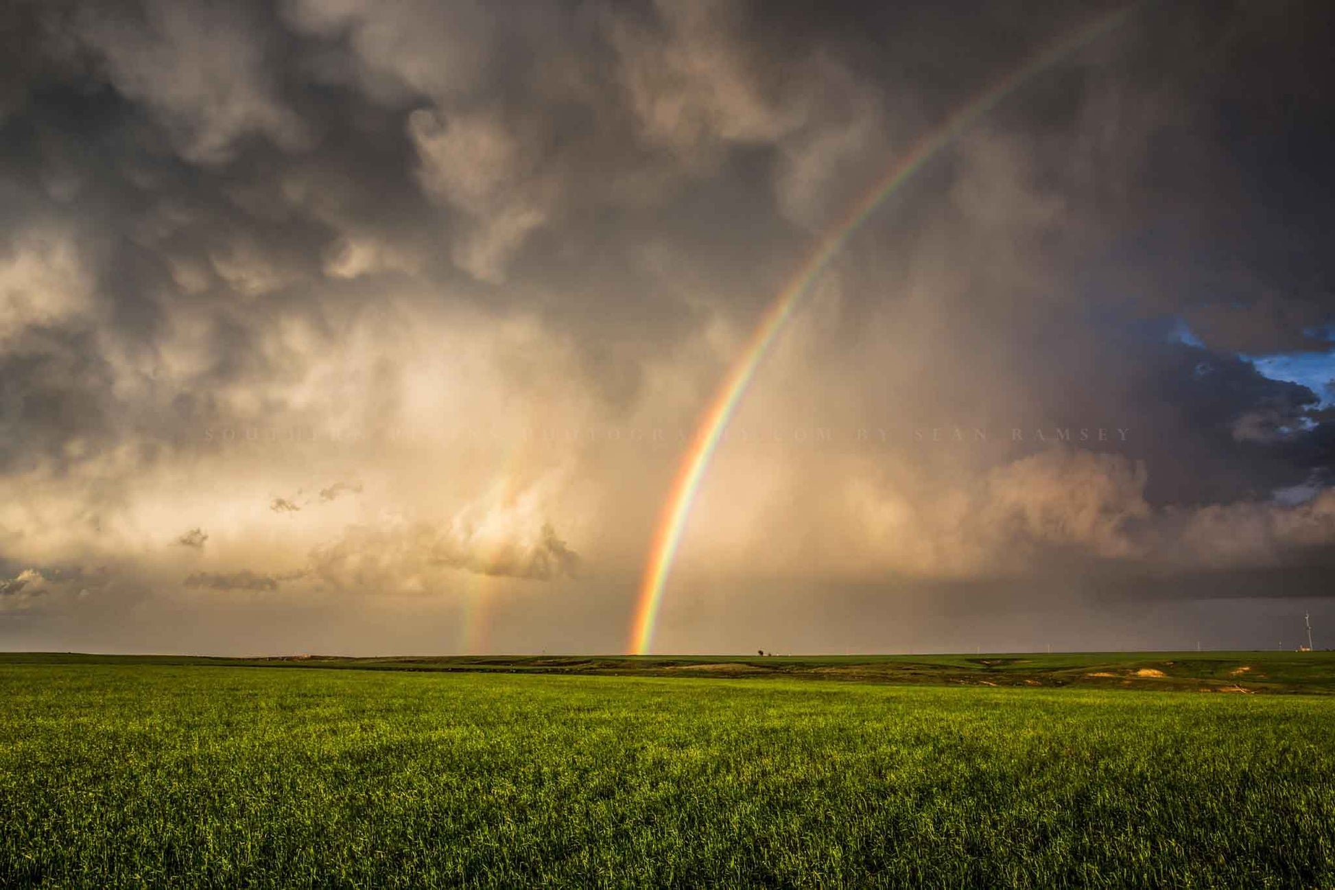 Nature photography print of a brilliant rainbow against a stormy sky ending in a green field on a spring day in Oklahoma by Sean Ramsey of Southern Plains Photography.