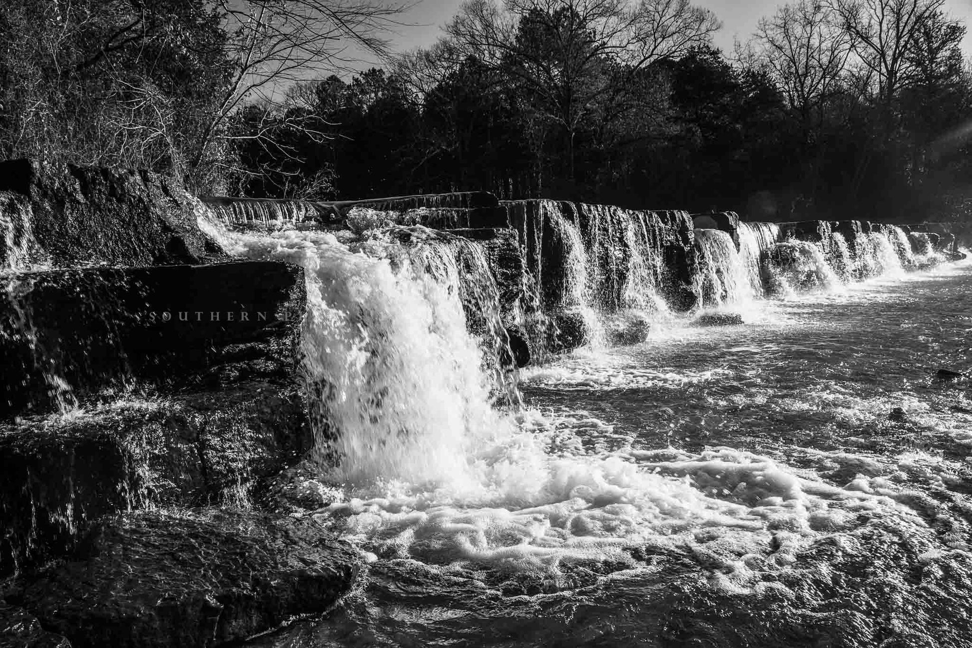 Black and white waterfall photography print of Mountain Fork Creek crossing over the rock ledges of Natural Dam Falls on a winter day in the Ozark Mountains of Arkansas by Sean Ramsey of Southern Plains Photography.