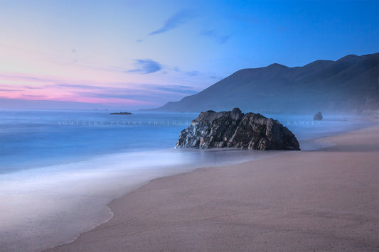 Coastal photography print of a large boulder on a beach as waves gently wash ashore with mountains in the background during a serene sunset in Big Sur, California by Sean Ramsey of Southern Plains Photography.