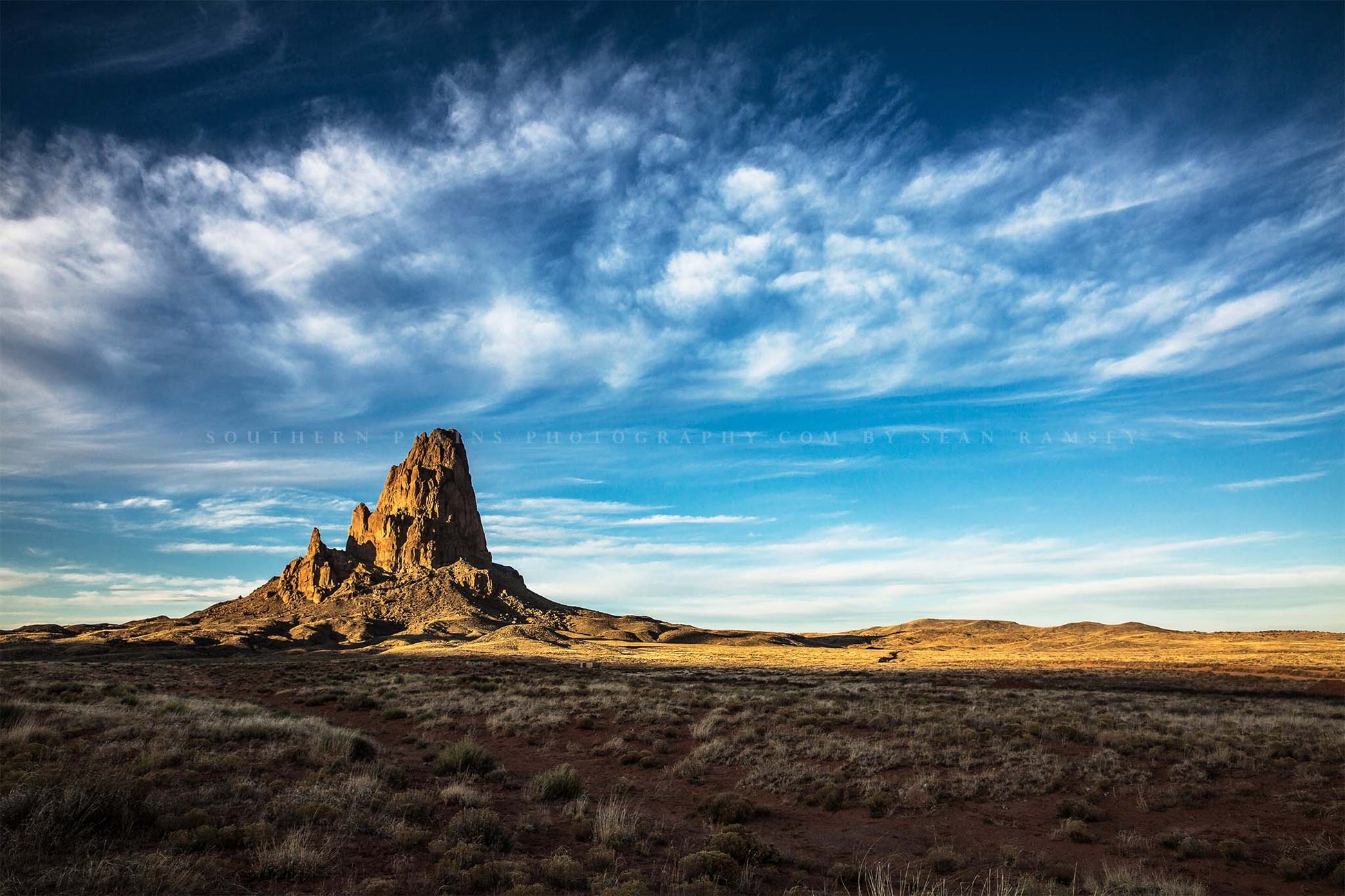Western landscape photography print of Agathla Peak drenched in evening sunlight under big blue sky full of wispy cirrus clouds by Sean Ramsey of Southern Plains Photography.