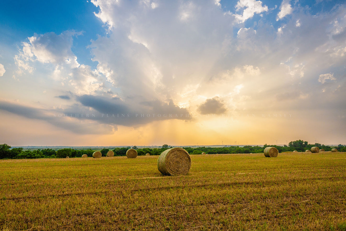 Country photography print of round hay bales under a stormy sky filled with golden sunlight on a spring day in Oklahoma by Sean Ramsey of Southern Plains Photography.