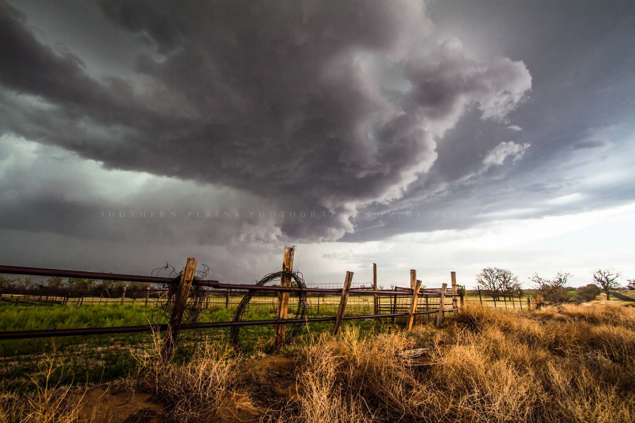 Western photography print of a thunderstorm advancing over rolled barbed wire leaning against a fence on a stormy spring day in Oklahoma by Sean Ramsey of Southern Plains Photography.